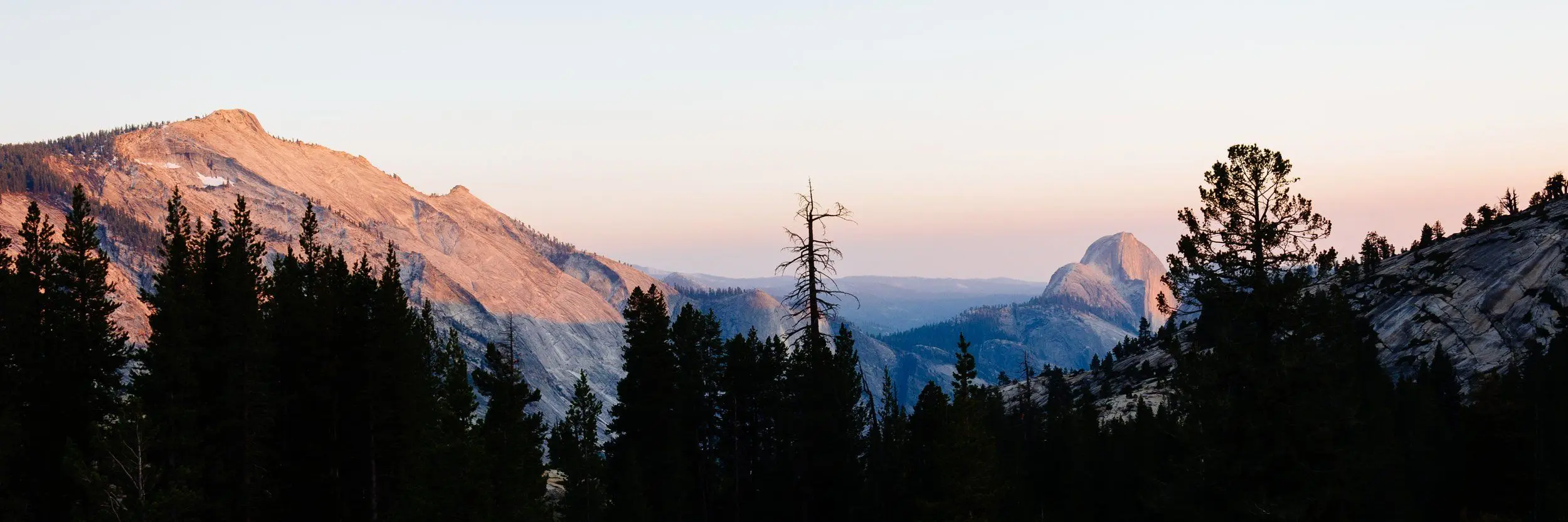 Wide shot at sunset including Half Dome in the distance with dark trees in the foreground.