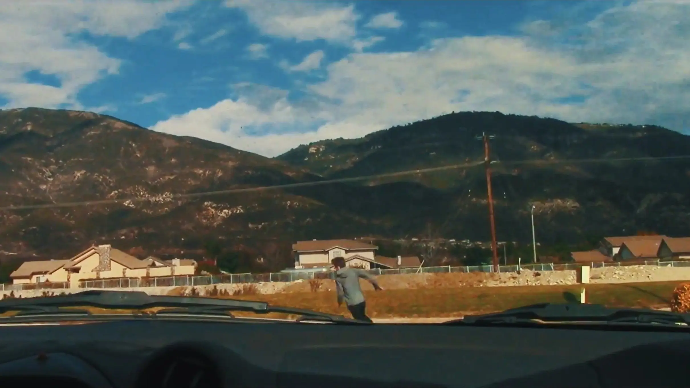 A man runs away from a car that we are in with a house and mountains in the background.