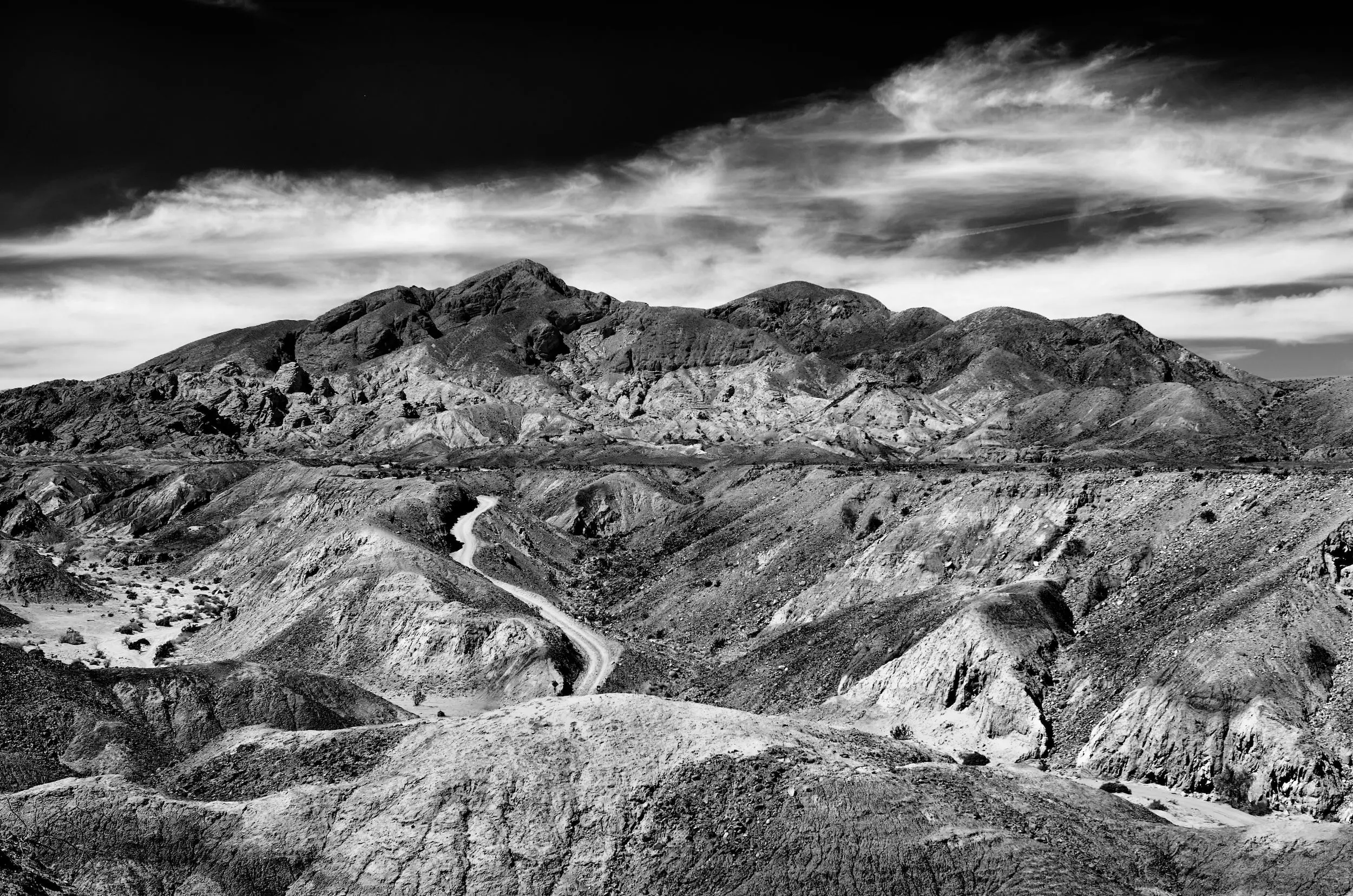 Black and white shot of a road cutting between rocky valleys, with mountains in the background and dramatic clouds in the black sky.