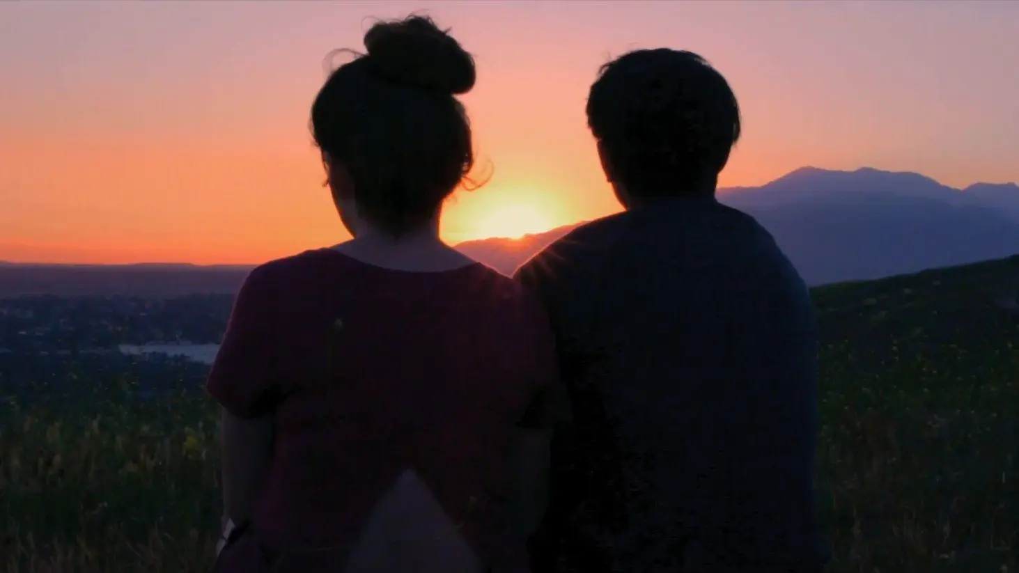 A couple sits together, backs to us, looking towards the setting sun.