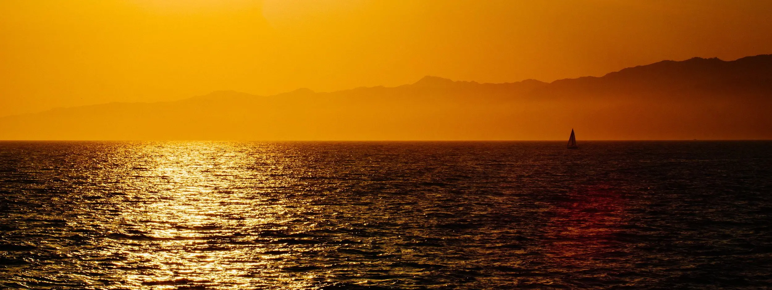 A sailboat near the horizon on the black ocean water, silhouetted against mountains. Golden hour.